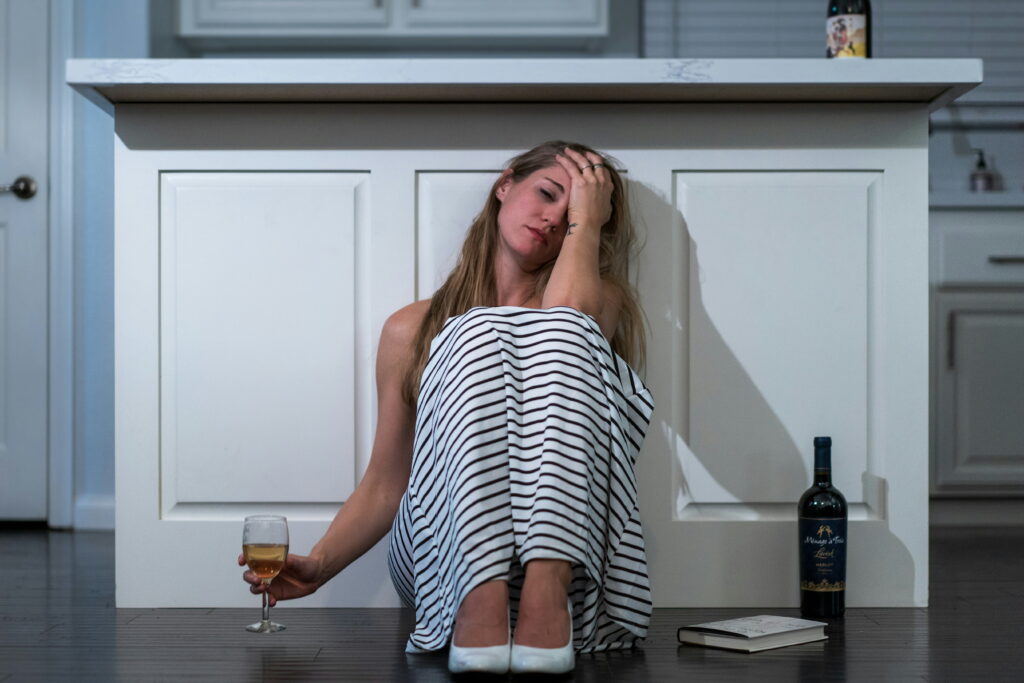 midlife woman on the kitchen floor looking depressed with wine glass and bottle